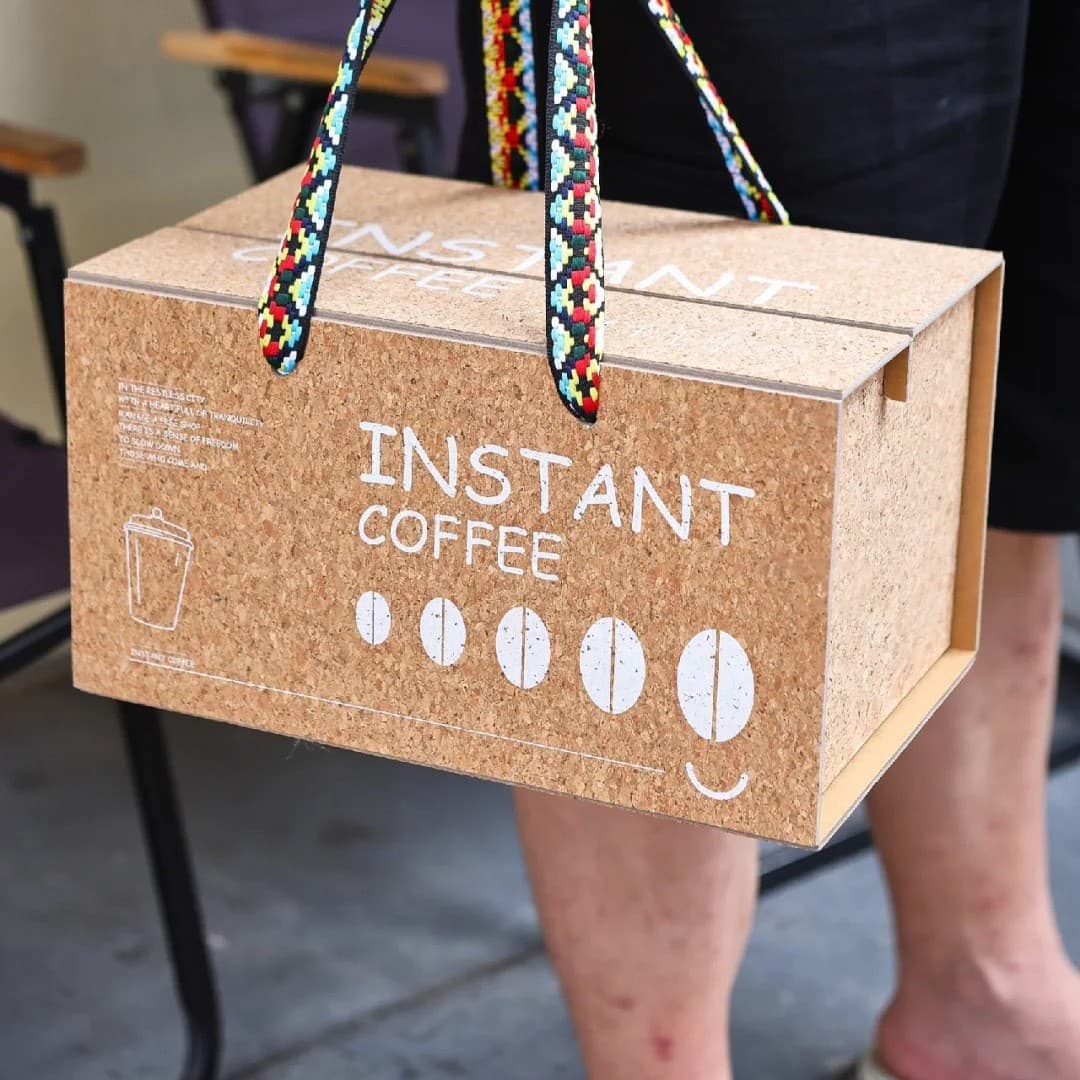 Person carrying a cork box labeled 'INSTANT COFFEE' with vibrant, multi-colored patterned woven straps.