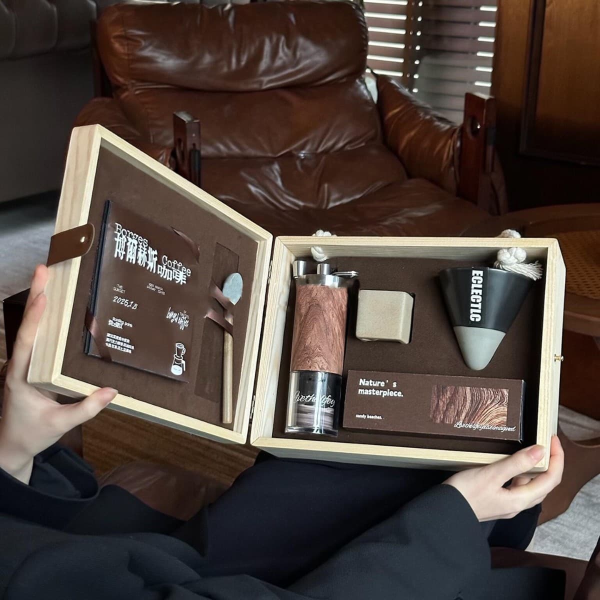 Hands holding an open wooden box containing a manual coffee grinder, ceramic dripper, and accessories.