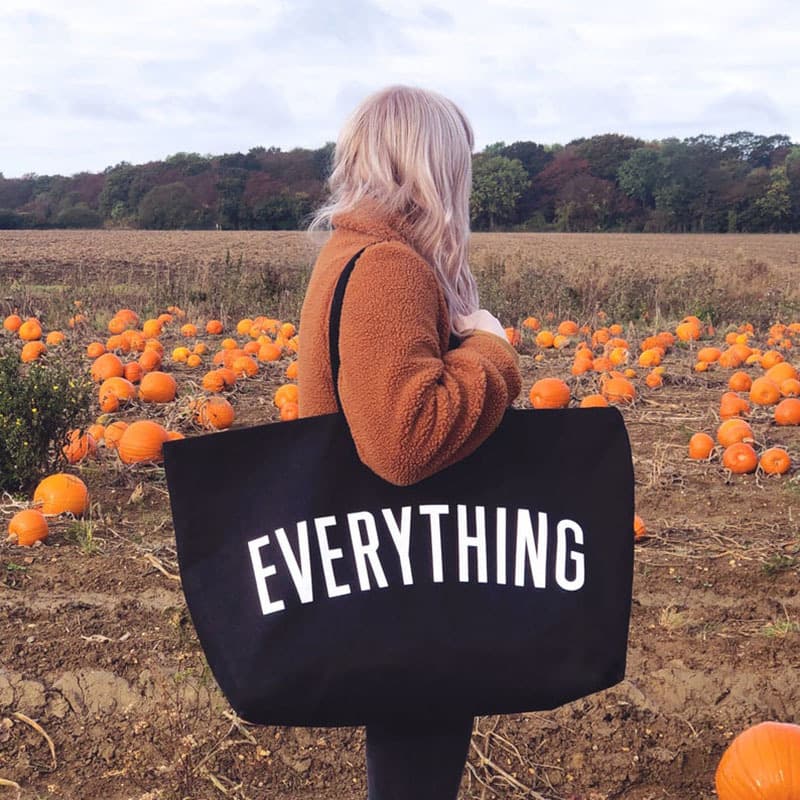 Person in brown jacket carrying a black 'EVERYTHING' tote bag in a pumpkin patch.